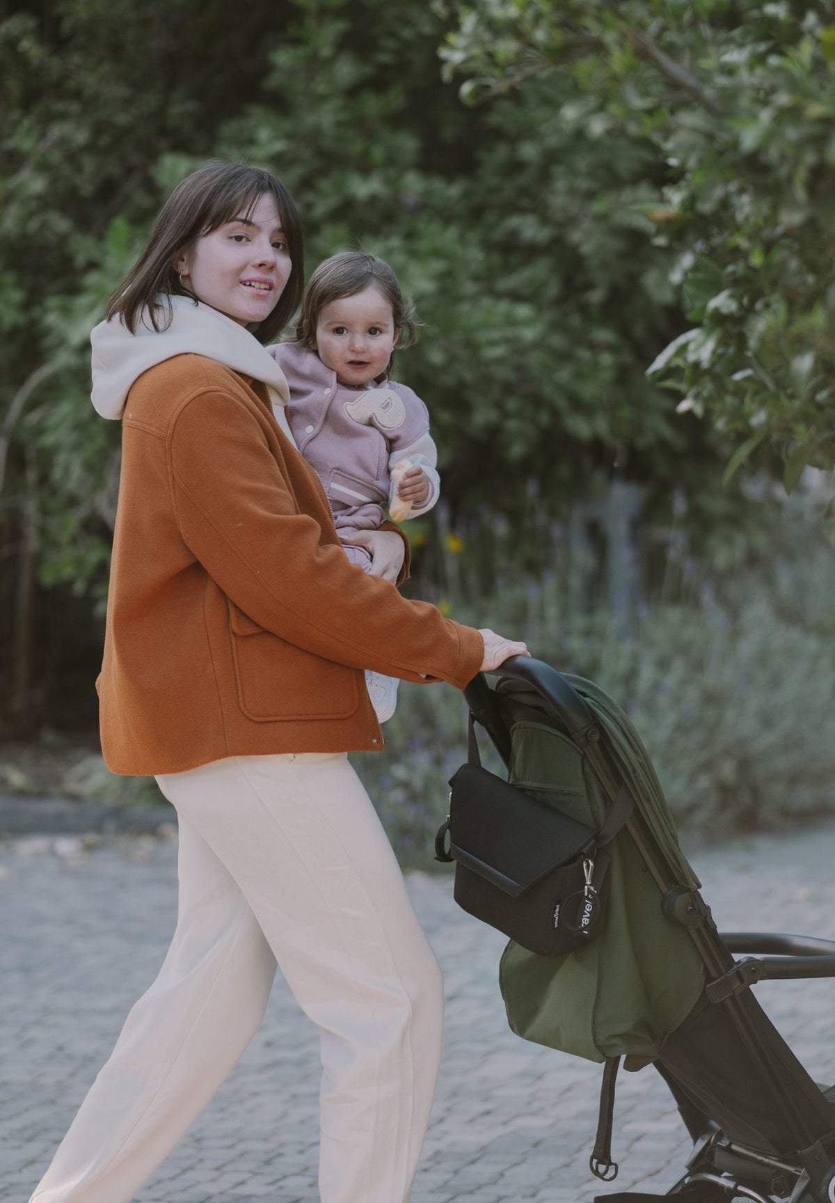 The image shows two women outdoors on a grassy area near a body of water under a clear sky. Each woman is pushing a stroller, with one toddler seated in each stroller. The children appear to be around one to two years old and are using pacifiers. The woman on the left has long hair and is wearing a light-colored vest over a white top. The woman on the right has her hair pulled back, wears sunglasses, a light blue shirt, dark pants, and a white sweater draped over her shoulders.