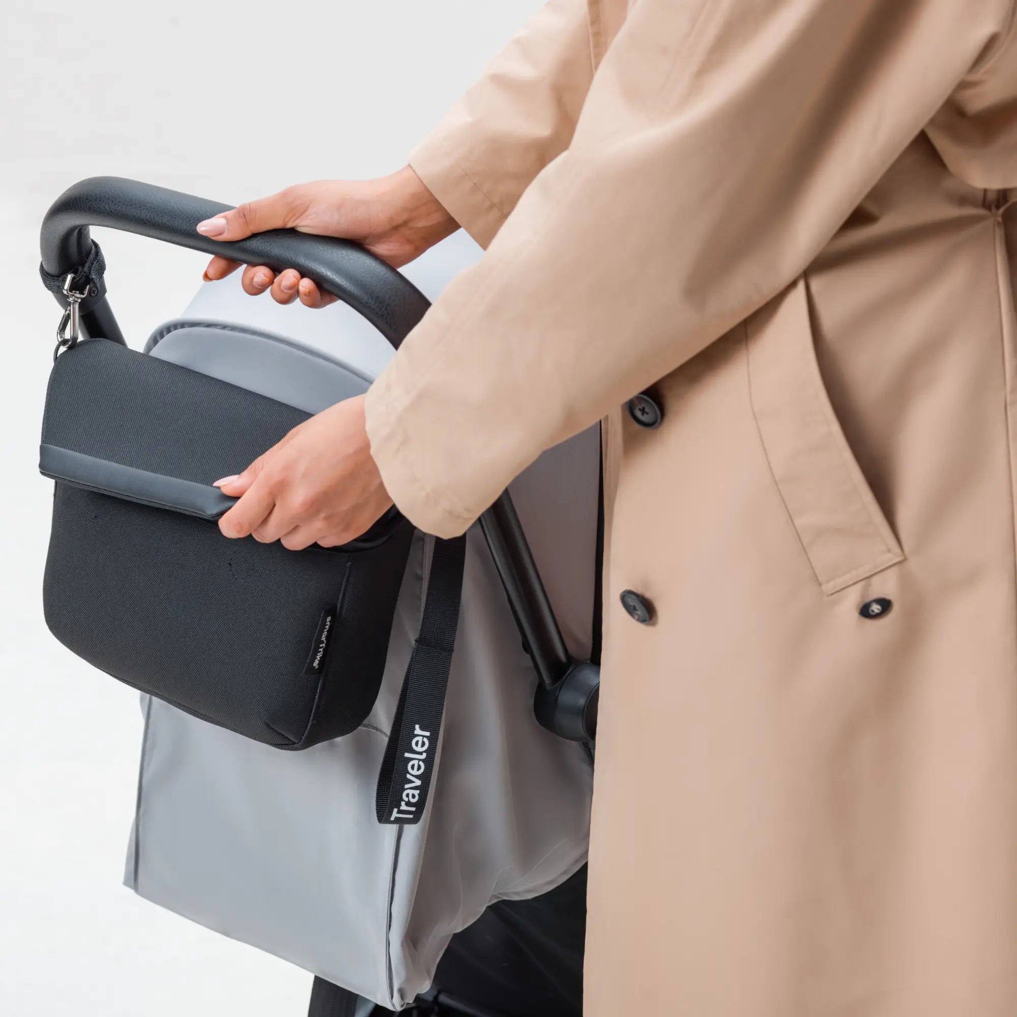 {"image_loaded": true, "load_issue": null, "description": "A person wearing a beige trench coat is holding the handle of a gray stroller. Attached to the stroller's handle is a black, rectangular bag with a flap closure and a small label. There is a black strap hanging from the stroller with the word 'Traveler' printed in white. The background is plain and light-colored.", "people": {"count": 1, "roles": ["caregiver or parent"], "visible_demographics": null, "attire": "beige trench coat", "pose_or_activity"