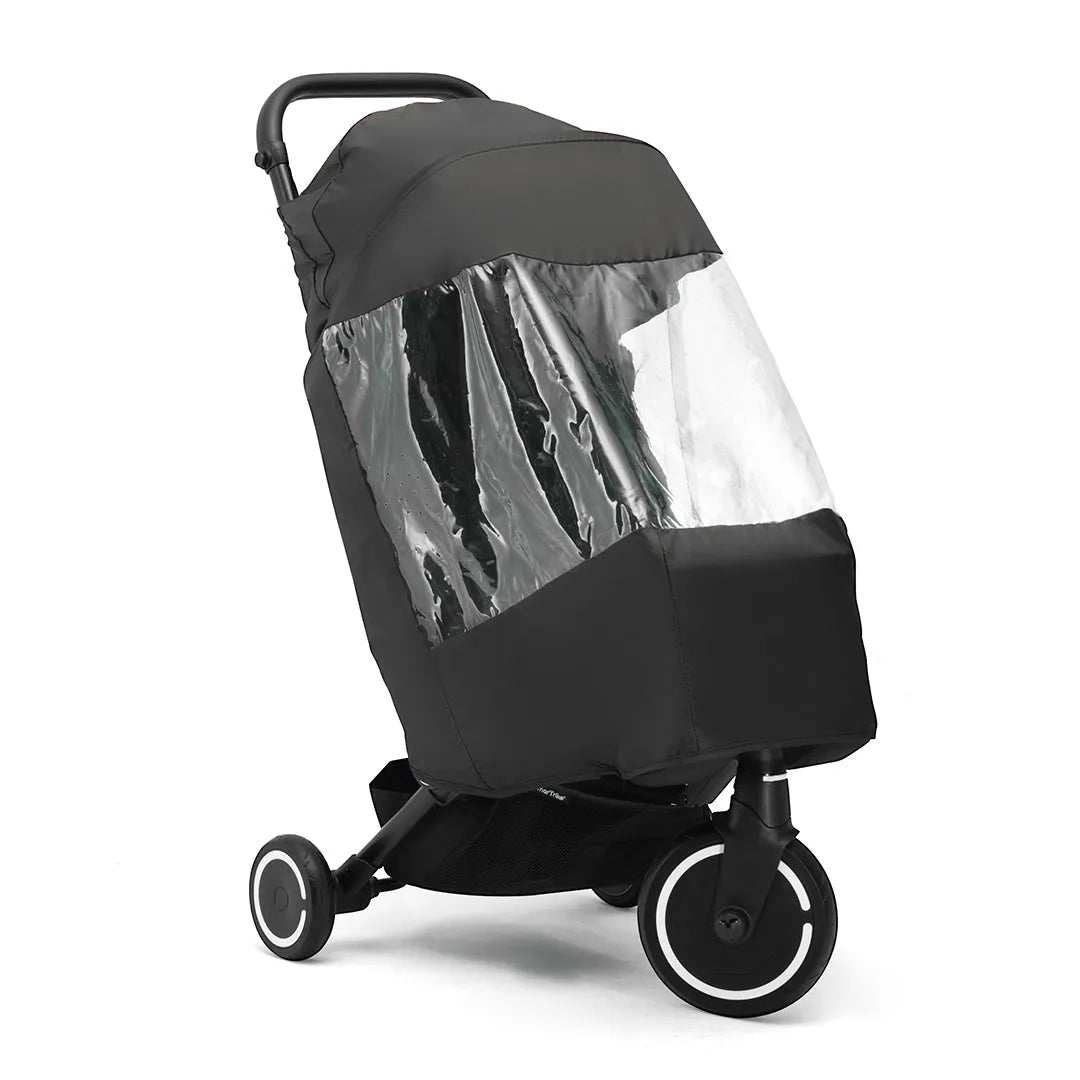 The image shows a black stroller with a protective rain cover on it. The cover has a clear plastic window on one side, allowing visibility inside the stroller. The stroller has three wheels: two smaller rear wheels and one larger front wheel with a white circular accent. The background is plain white, and the lighting is even and neutral, eliminating shadows.
